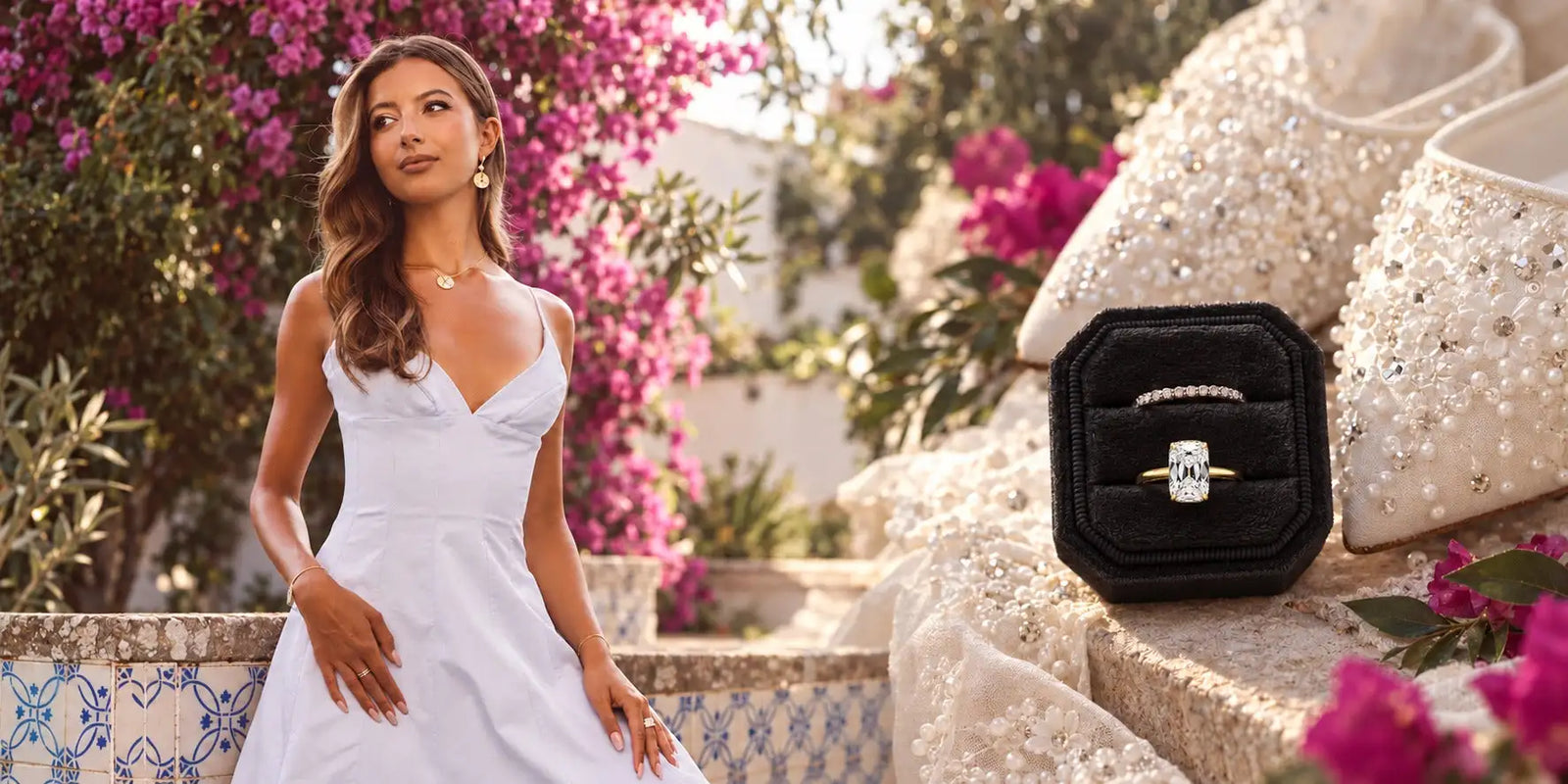 diamondboi hero: a model in a white dress in a garden of pink bougainvillea beside a black velvet ring box holding a cushion-cut diamond engagement ring and a diamond band
