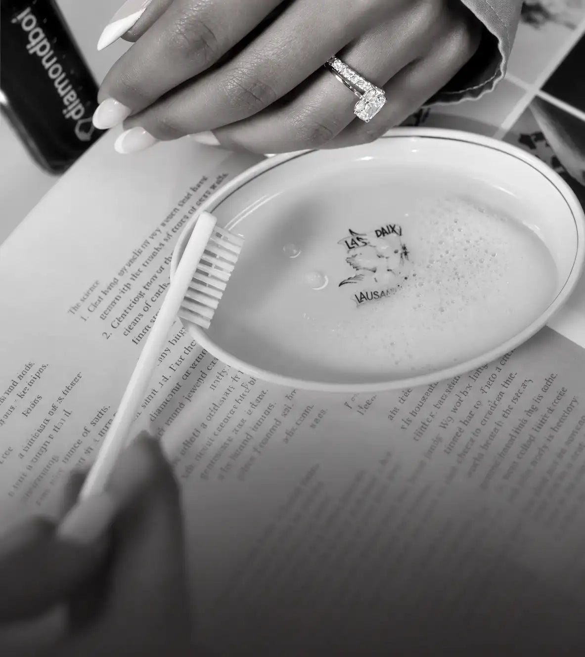 A hand with a diamond ring holds a white toothbrush over a small, round dish containing soapy water and faintly visible text.