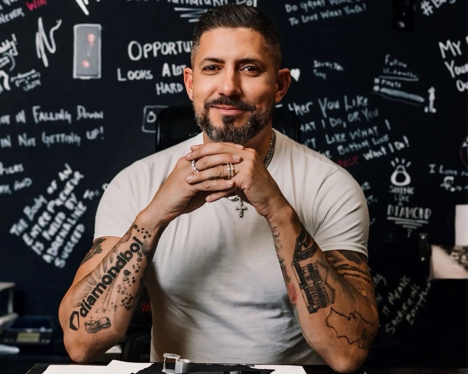 Man with dark hair and beard wearing a white t-shirt, visible tattoos on both arms, and multiple rings.