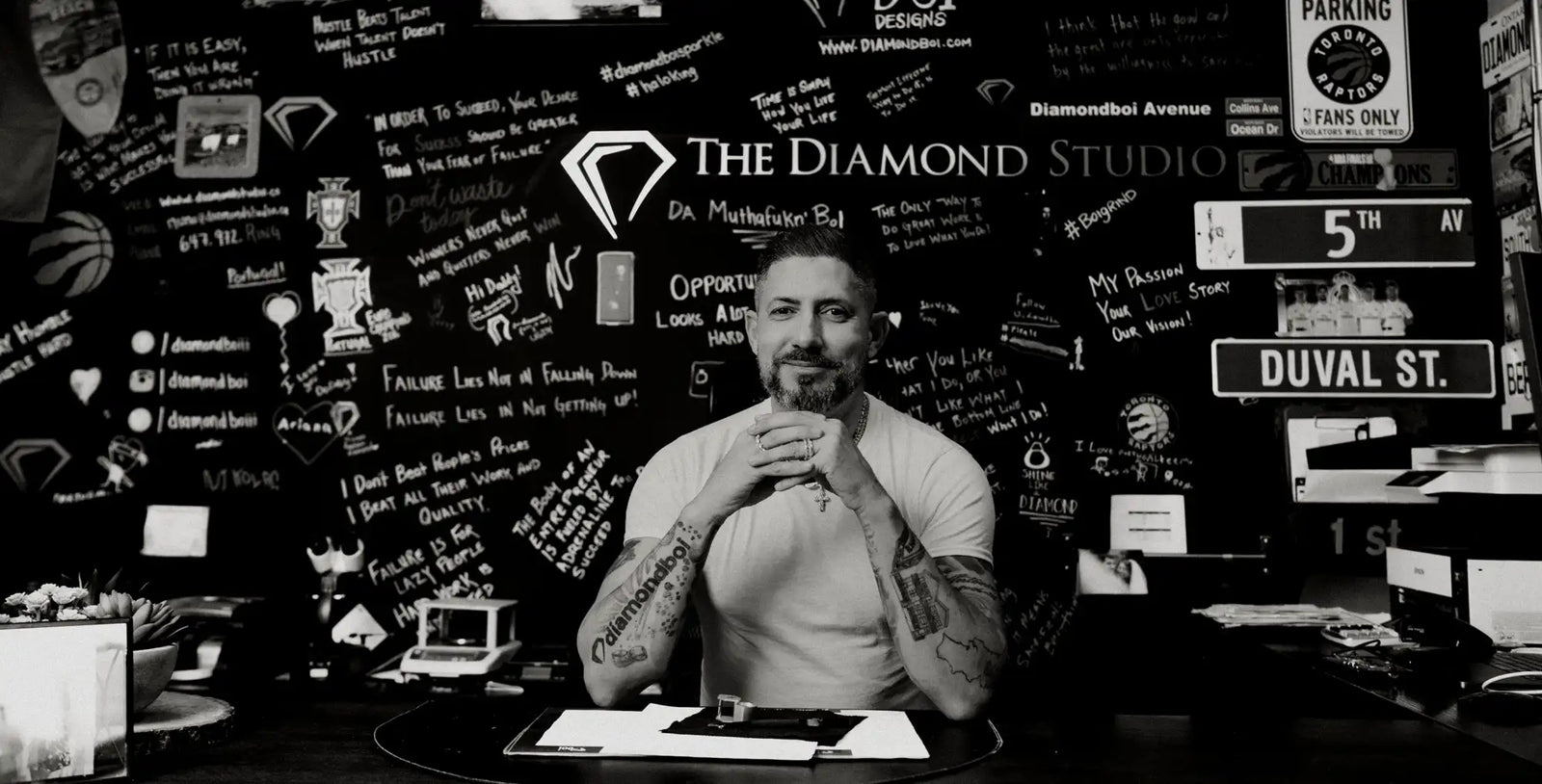 Man with tattooed arms wearing a white t-shirt sitting at a desk in front of a wall covered in handwritten notes and street signs