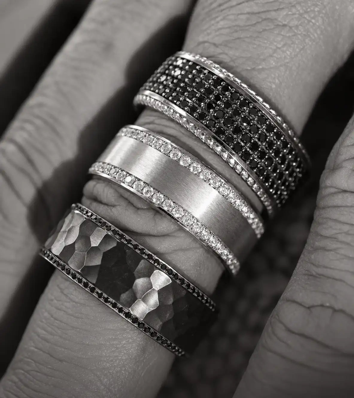 Three stacked men’s rings in silver-toned metal, featuring intricate designs with black and clear gemstones.